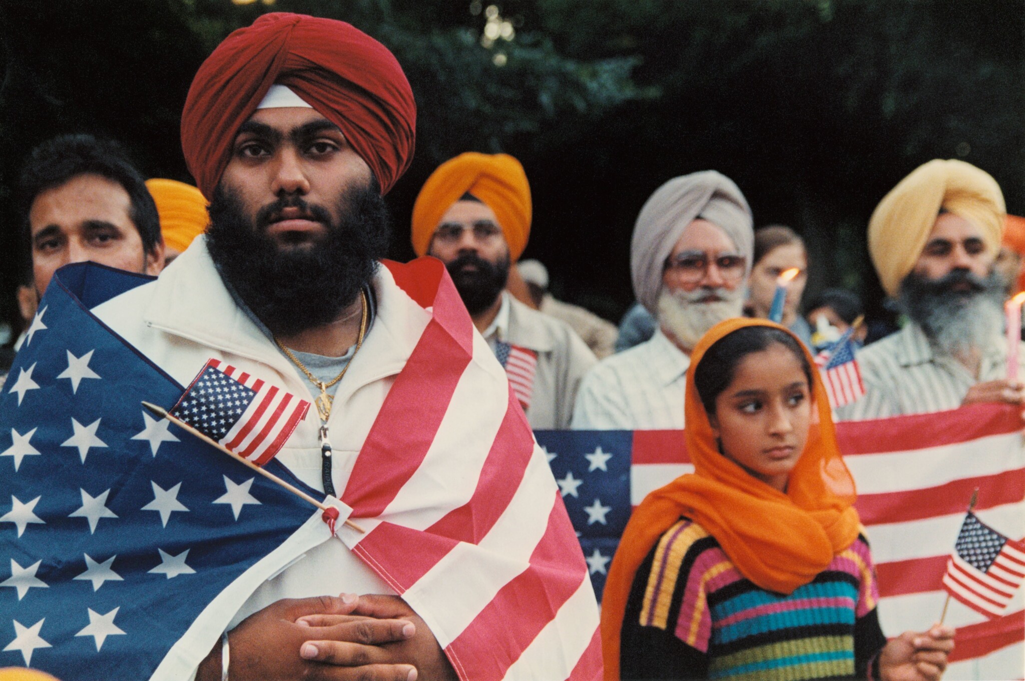 A Sikh American man stands with an American flag wrapped around his shoulders, with more Sikh men and a young Sikh woman in the background.