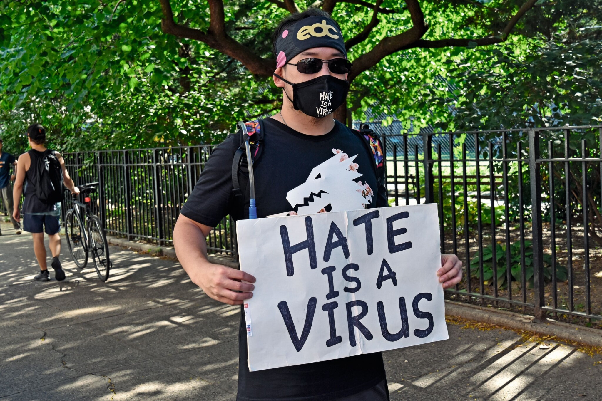 An Asian American man in a black shirt holds a sign and wears a face mask that say "Hate is a virus."