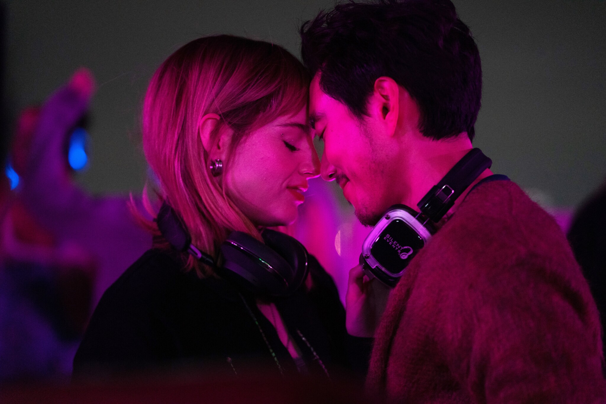 Lucy Boynton and Justin H. Min in "The Greatest Hits" stand with their foreheads together, with headphones around their necks.