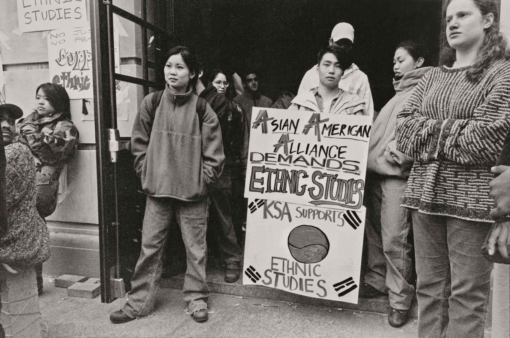 A young Asian Man holds signs demanding ethnic studies, surrounded by other young people of different races.