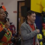 A Black woman and Asian man stand with a microphone in their hands with balloons in the background.