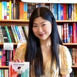 Author Kyla Zhao rests a hand on the book "Valley Verified" in front of her and a bookshelf filled with books in the background.