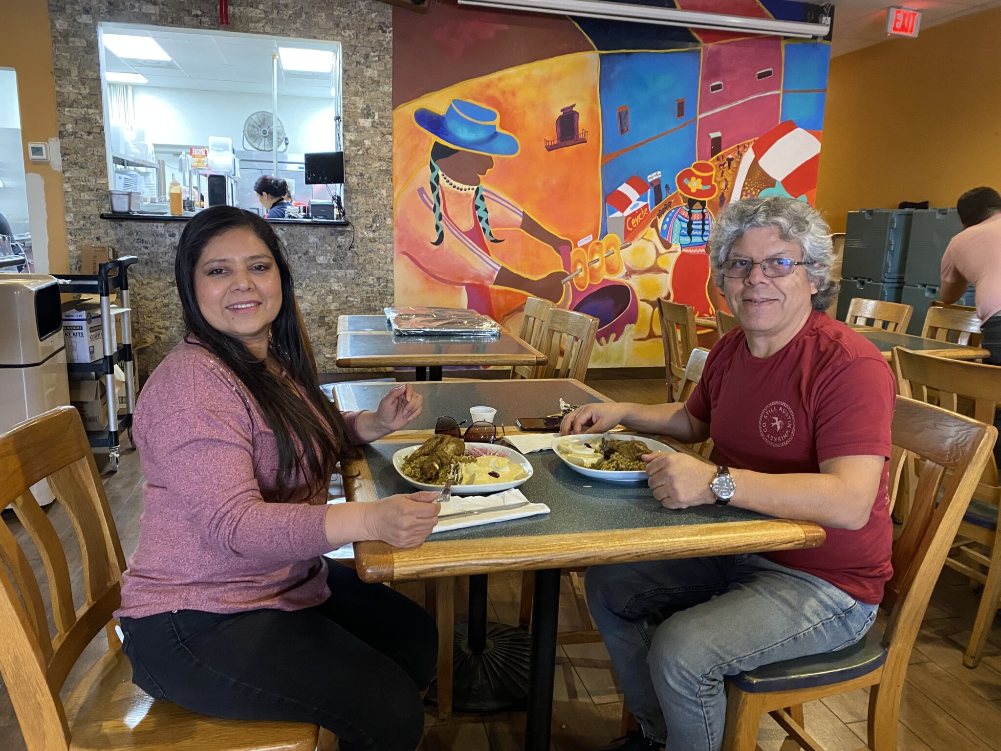 A Latino man and woman sit at a restaurant table, with plates of food in front of them and a colorful mural in the background.