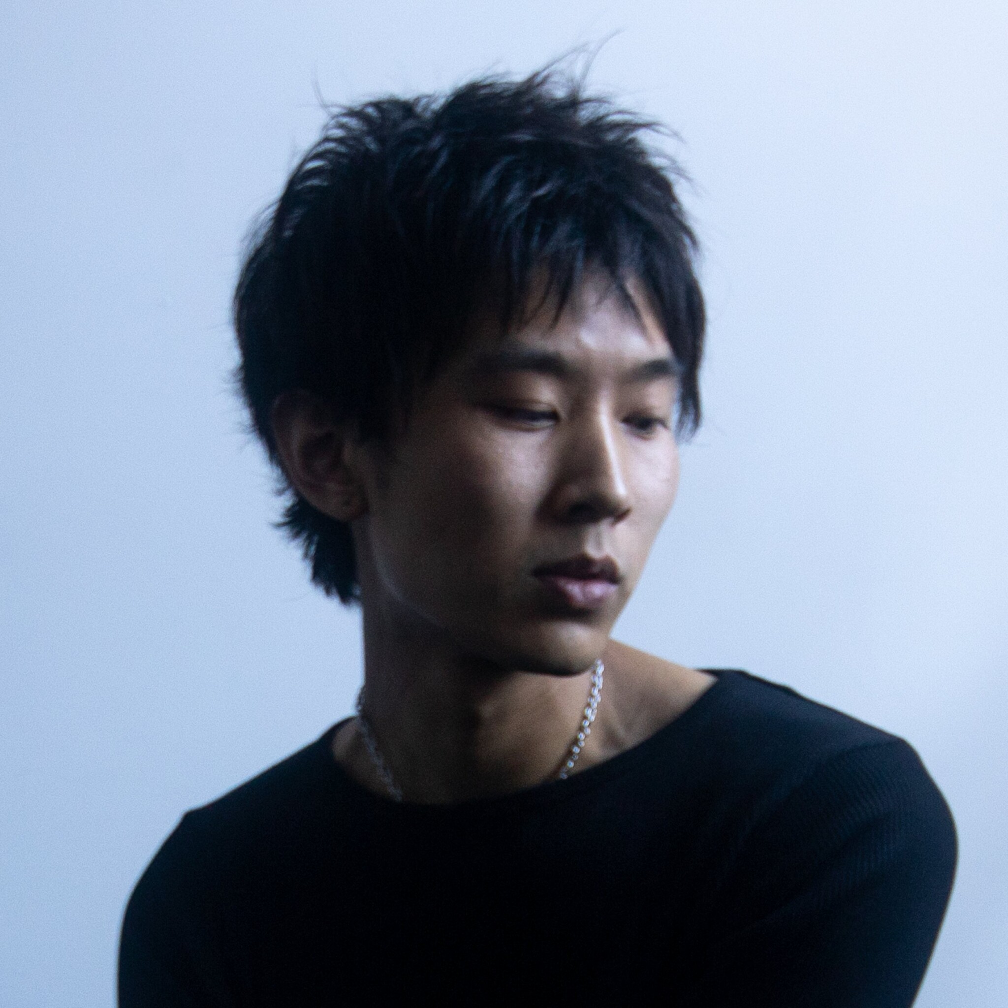 A young Asian man with black hair, in a black top, looks down and to the right, against a light blue background.